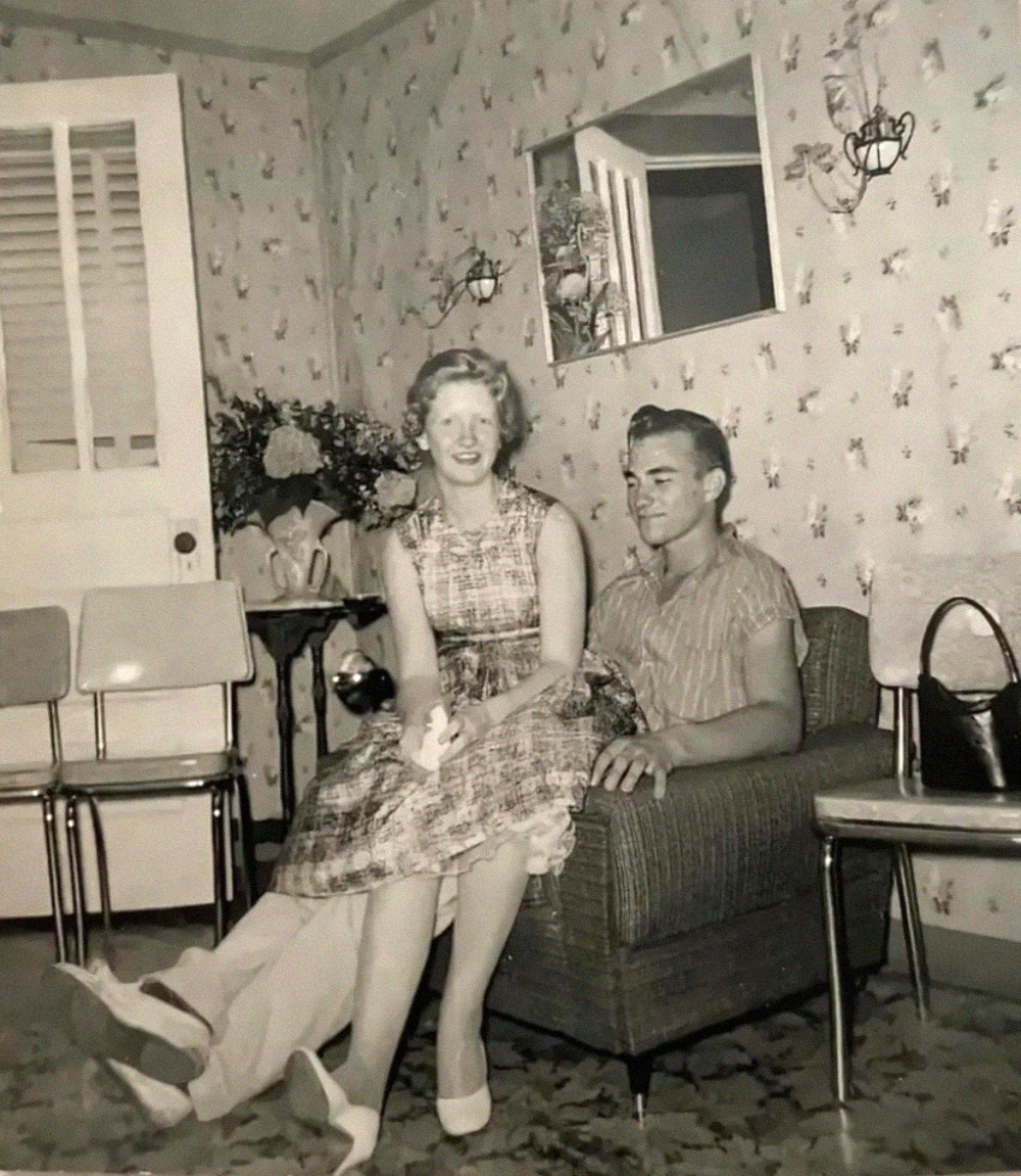 A young couple sits together in a living room, with the woman perched on the arm of an upholstered chair beside a man.
