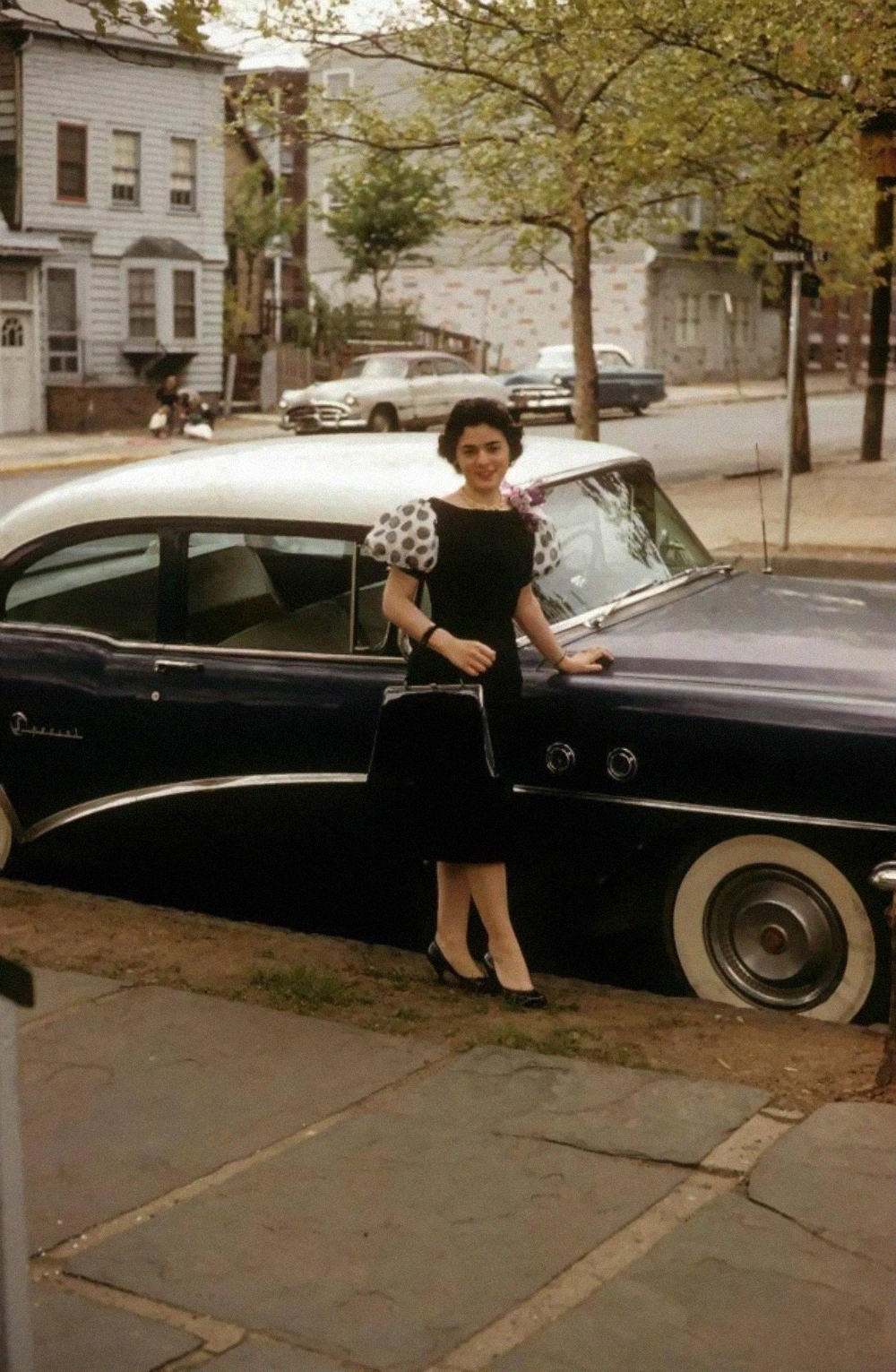A woman in a black dress with white polka-dot sleeves stands on a sidewalk next to a dark blue vintage car.