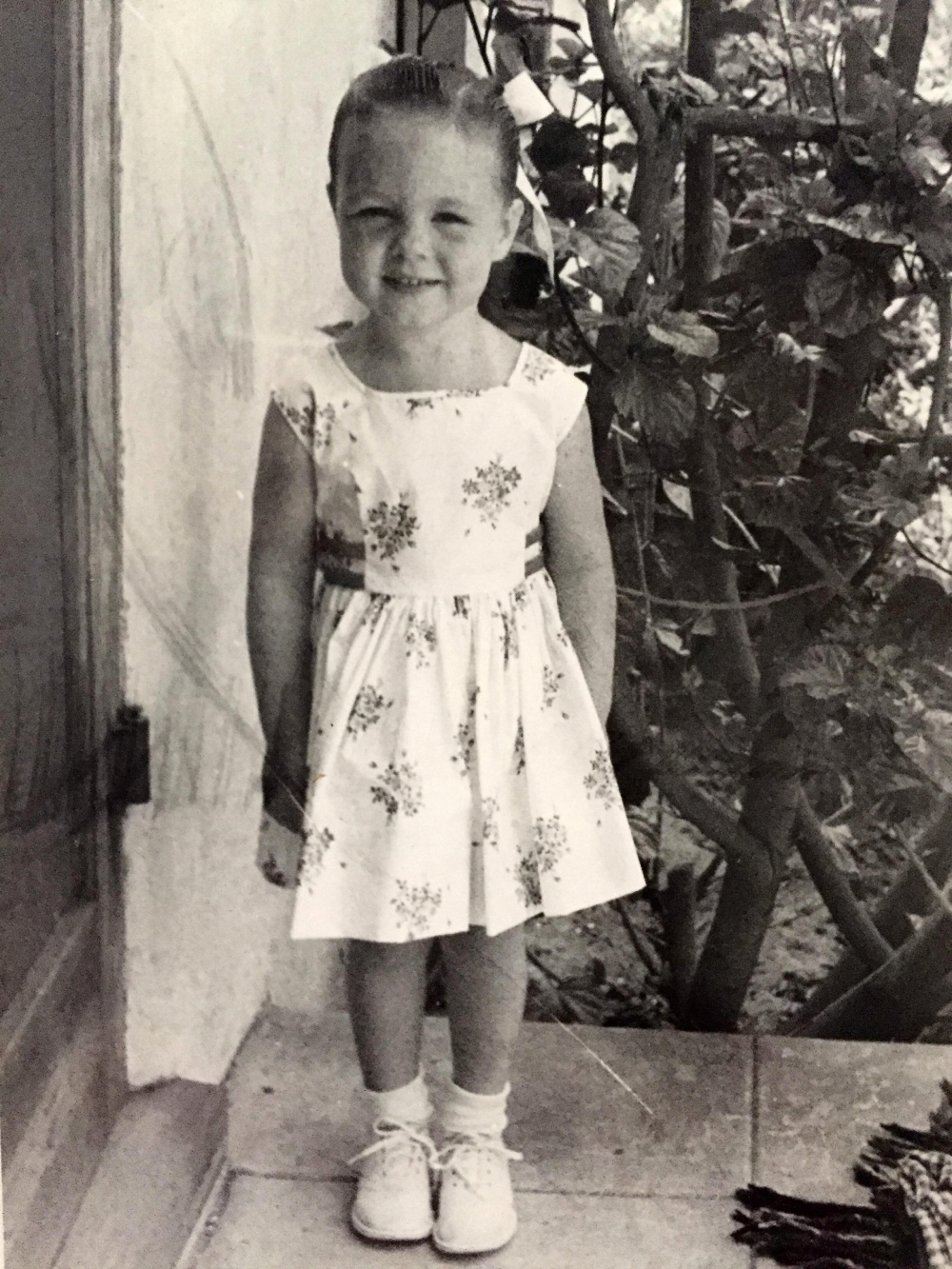 A young girl with a hair bow stands outdoors wearing a floral dress, socks, and tied shoes.