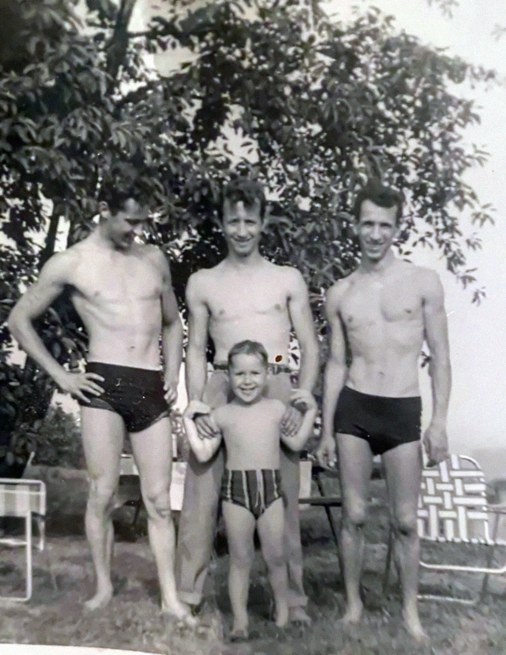 Three men in swim trunks and a young child stand outdoors on grass, smiling for a black-and-white photo. Two men hold the child’s hands. Trees and outdoor chairs are visible in the background.