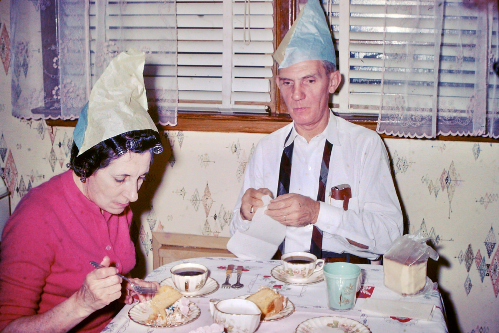 A woman in a pink sweater and a man in a white shirt and tie, both wearing paper party hats, sit at a table eating cake and drinking coffee.