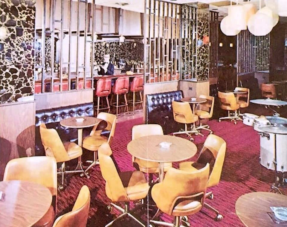 A mid-century restaurant interior with several round tables surrounded by swivel chairs, and a bar with red stools in the background.