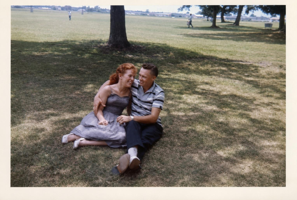 A smiling couple sits close together on the grass under a tree in a sunny park, holding hands. The woman wears a dress and the man a striped shirt. Other people and trees are visible in the background.