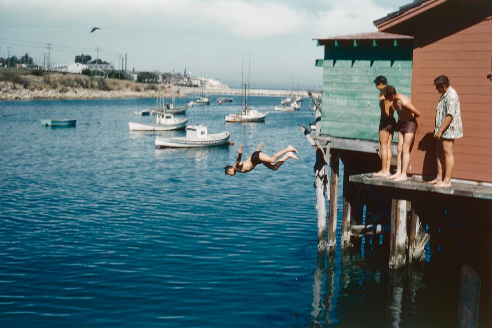 A person dives off a dock into the water while three others watch. Boats float nearby, and colorful wooden buildings are on the right. The sky is clear and it appears to be a sunny day.