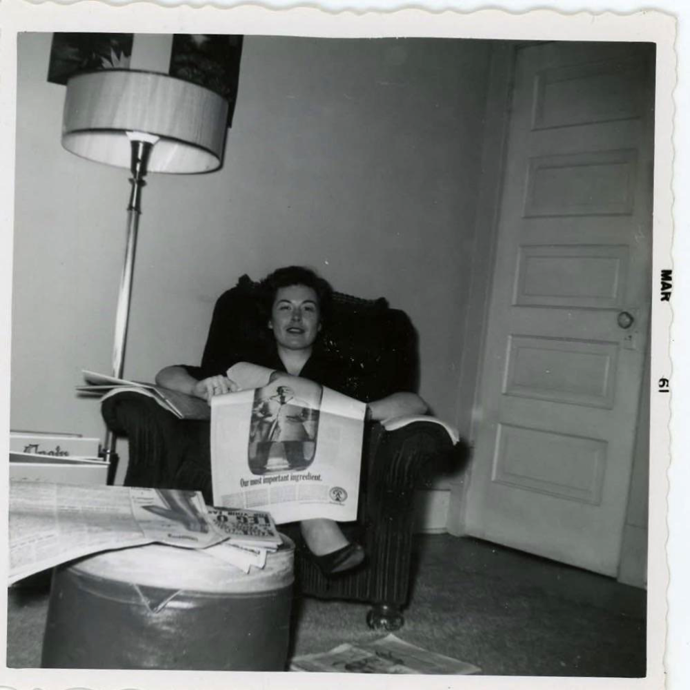 A woman sits in an armchair holding a newspaper, with a lamp and door behind her. Newspapers are scattered on a nearby table and floor. The photo appears to be from the 1960s.