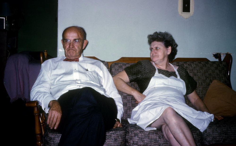 An older man in a white shirt and bow tie sits on a couch next to an older woman in a dark dress and white apron. Both look away from each other, with neutral or stern expressions, in a modest living room.