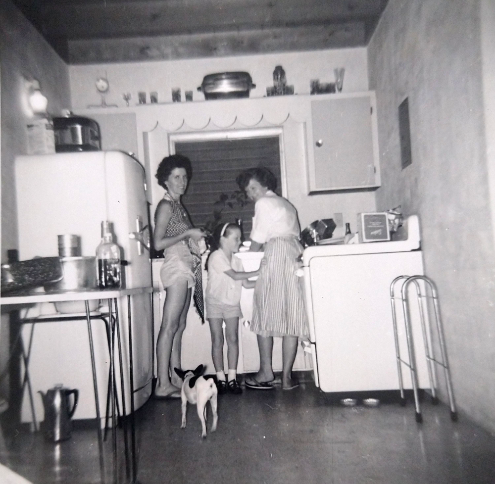 Three people and a small dog stand in a vintage kitchen. Two women work at the stove, while a young girl stands between them. A refrigerator, table, and kitchenware are visible. The photo is black and white.