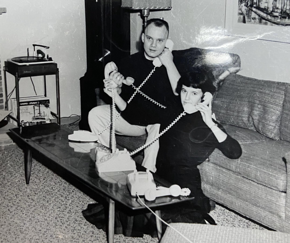 A man and woman sit in a living room, each holding a corded telephone. The man sits on the couch, the woman sits on the floor. A record player and lamp are visible in the background.
