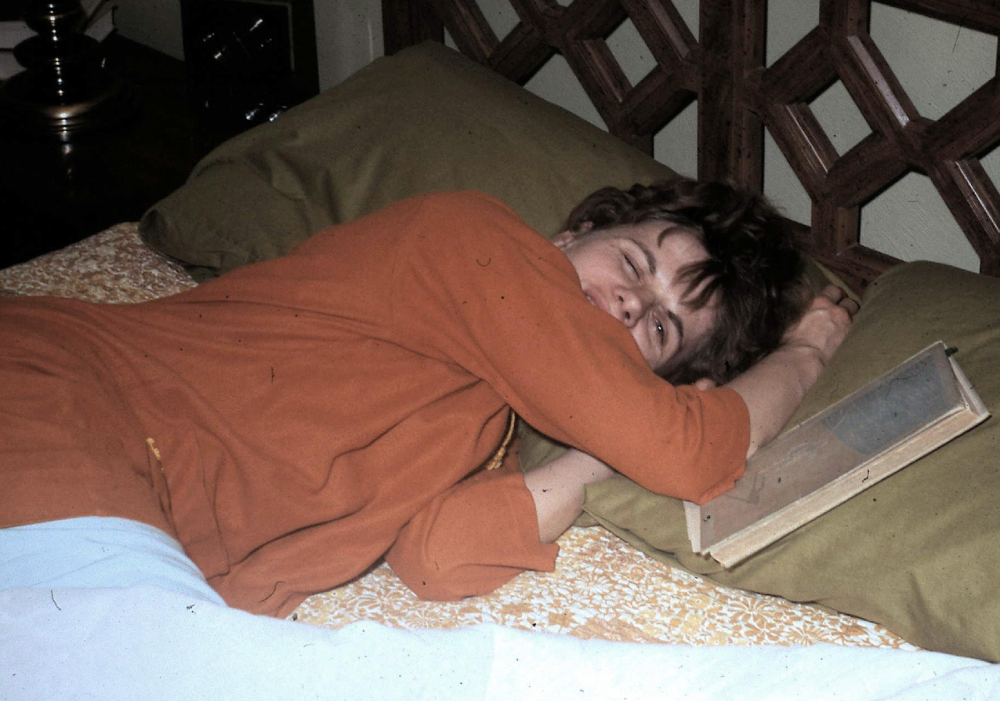 A person lies on a bed with green pillows, wearing an orange top, partially covering their face with an arm, and resting next to an open book.
