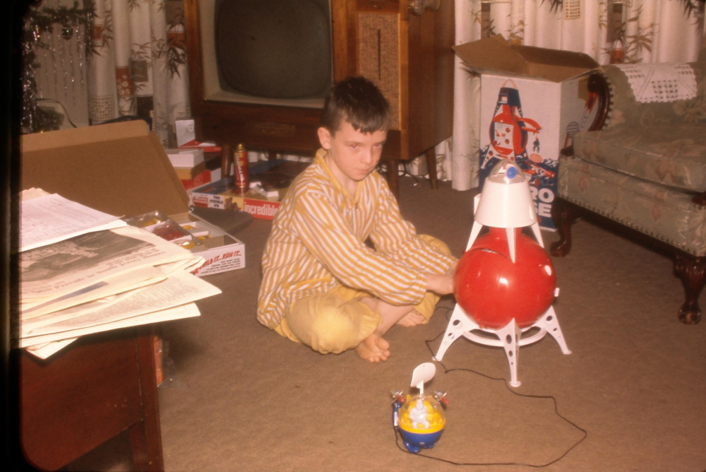 A boy in yellow striped pajamas sits on the floor playing with a red toy rocket. Behind him are a TV, a couch, and an open box. Papers and other toys are scattered around the living room.