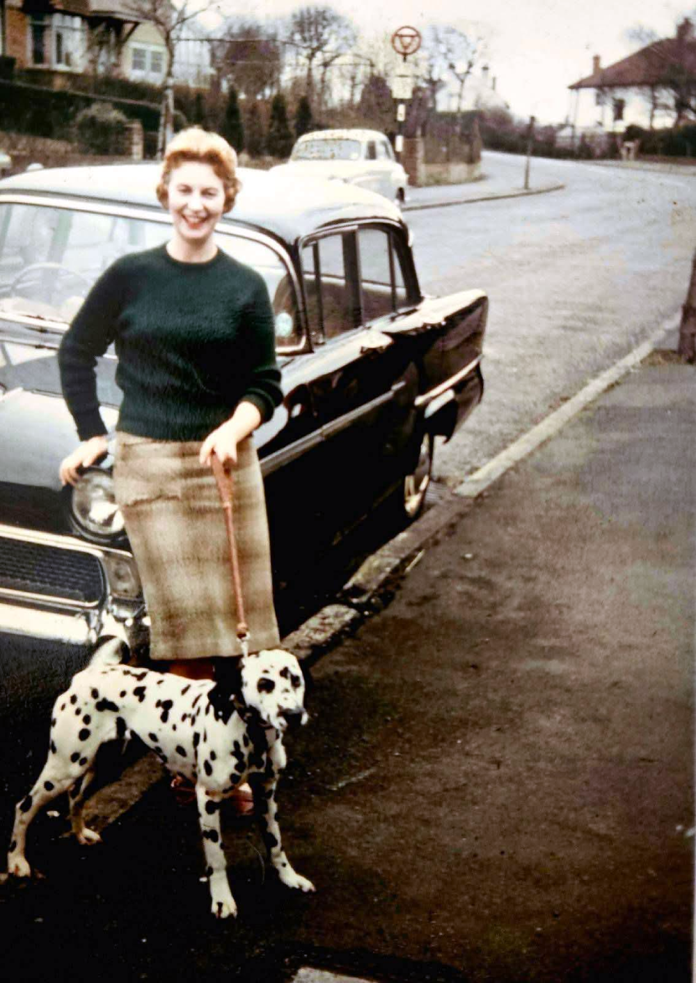 A woman in a green sweater and plaid skirt stands on a sidewalk beside a black vintage car, holding the leash of a Dalmatian dog. A street and houses appear in the background.