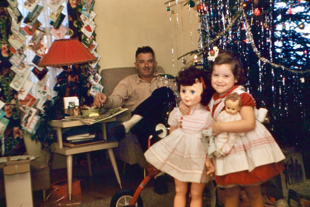 Two young girls in dresses stand by a Christmas tree holding dolls, while a man sits in an armchair nearby. Greeting cards are displayed on the wall, and gifts and decorations fill the cozy room.