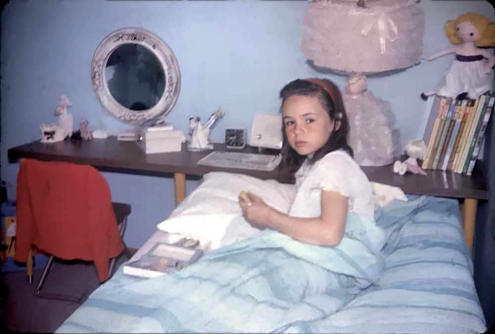 A young girl with dark hair sits in bed under a blue-striped blanket, holding something. Behind her is a desk with books, a lamp, a mirror, and a doll, with a red jacket draped over a chair.