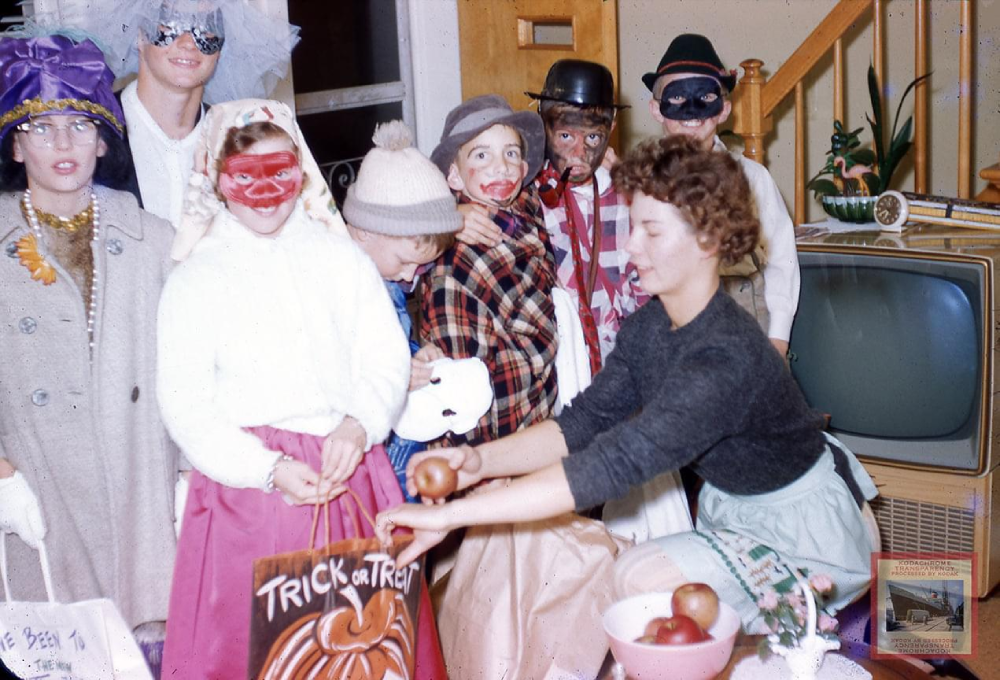 A group of children in Halloween costumes and masks stand indoors as a woman hands out apples. A vintage TV and staircase are visible in the background. A “Trick or Treat” bag is held open in the foreground.