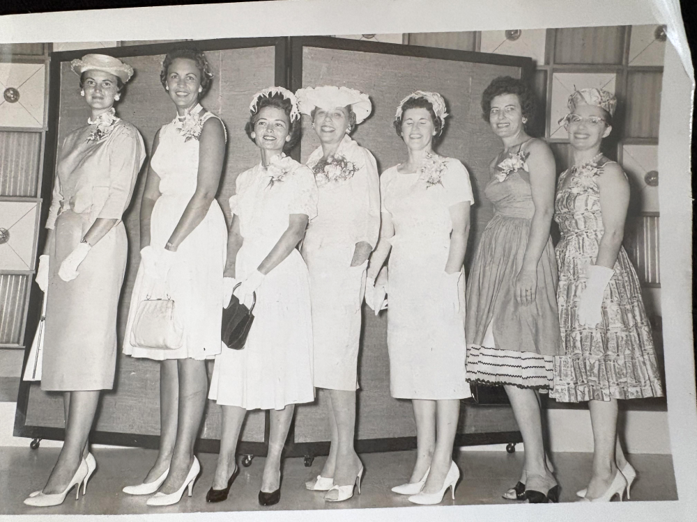 Six women stand side by side, dressed in elegant mid-20th century attire with hats, gloves, and corsages, smiling for the camera in a black-and-white setting.