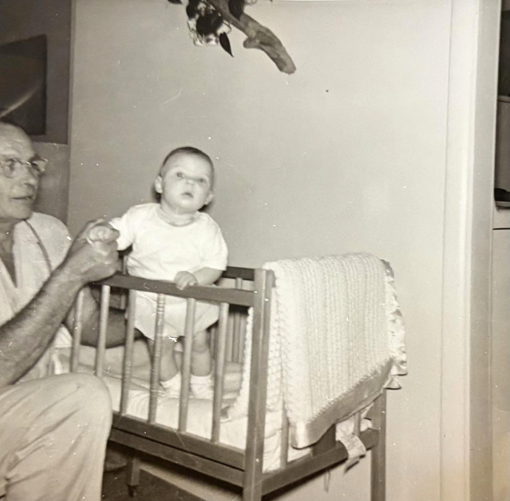A man holds a baby who is sitting in a wooden crib with a blanket draped on the side, in a black-and-white photo.