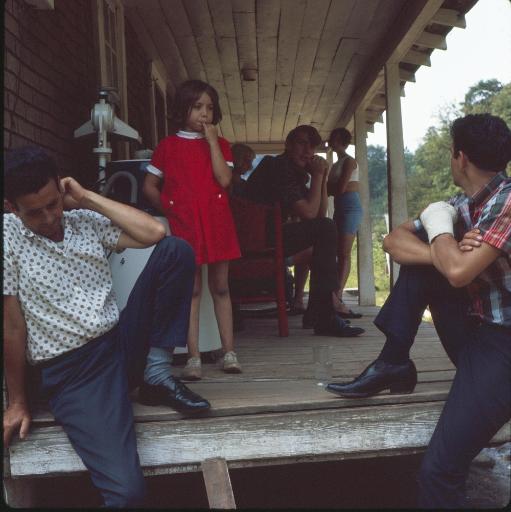 A girl in a red dress stands on a wooden porch with several adults sitting and talking around her. Two men sit separately at the edges, and one has a bandaged hand. The mood appears contemplative.