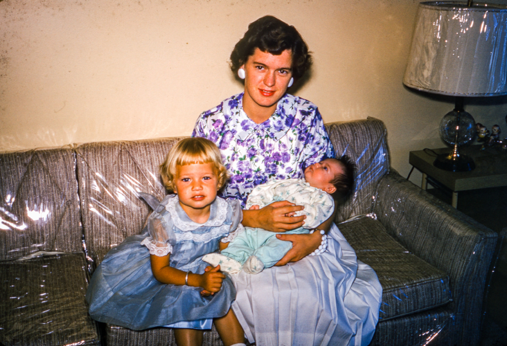A woman in a floral blouse sits on a couch holding a baby, with a young girl in a blue dress beside her. A lamp and clock are on a table nearby. The couch is covered with plastic.