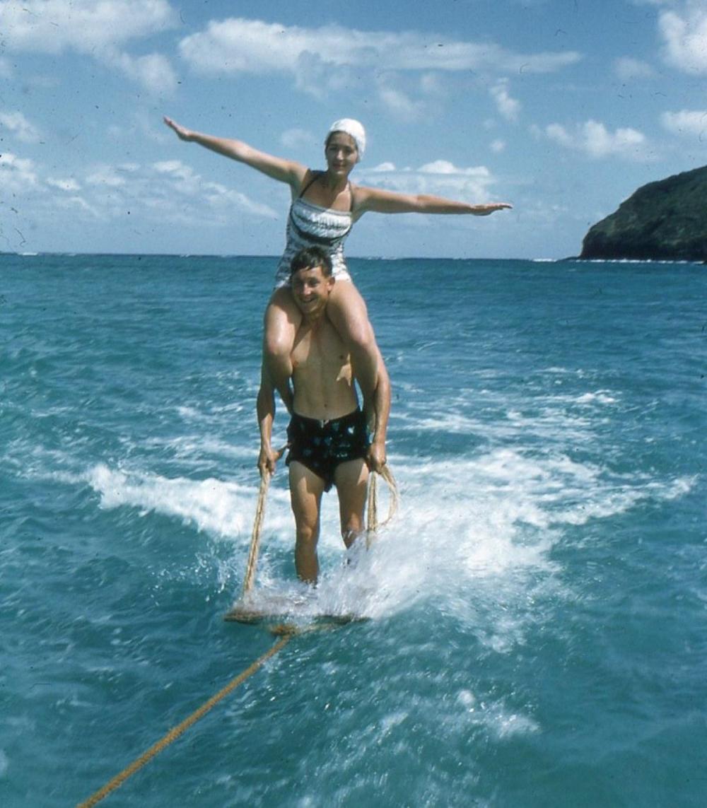 A woman balancing on a man’s shoulders while both ride a wooden board, being towed across blue ocean water on a sunny day with a small island in the background.