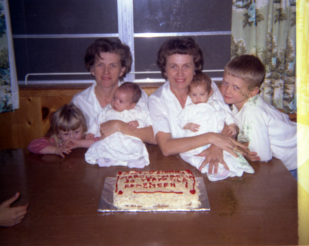 Two women sit at a table with four children, including two infants held in their laps, behind a rectangular sheet cake with red icing lettering.