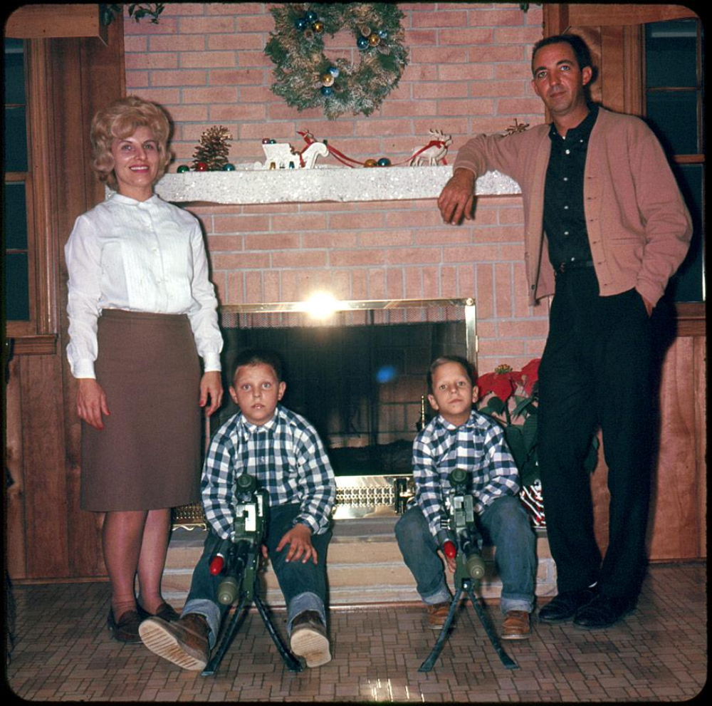 A family stands in front of a brick fireplace, with two boys sitting in front holding model rocket launchers.