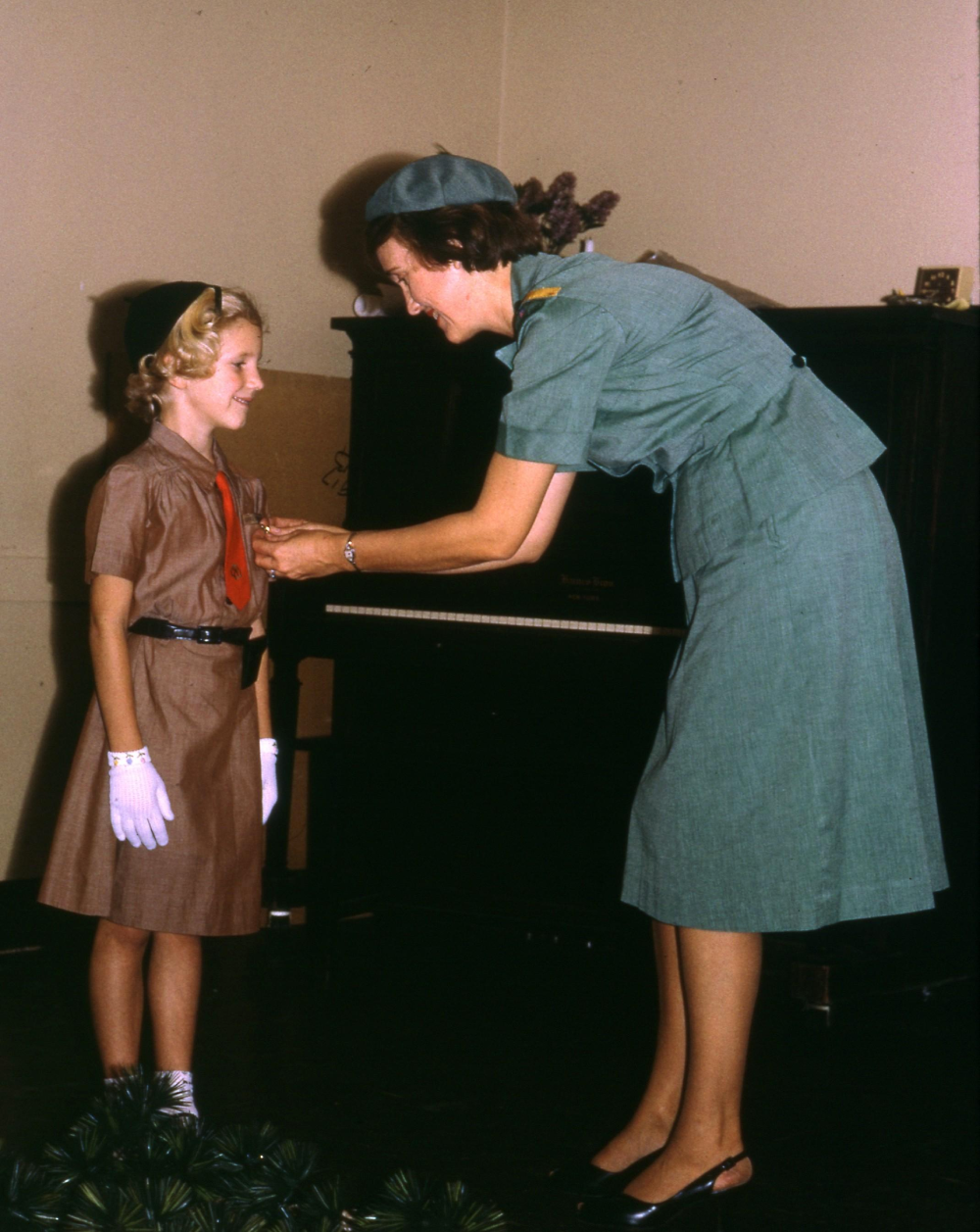 A Girl Scout leader in a blue uniform pins a badge on a young Brownie in a brown dress and white gloves. A piano stands in the background. Vintage photo.
