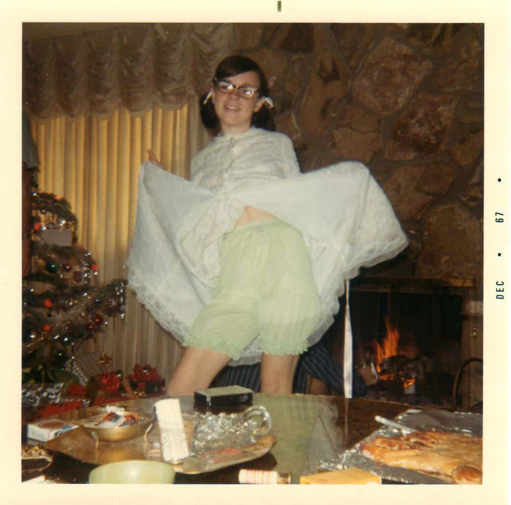 A person with glasses stands in front of a fireplace and Christmas tree, holding up the hem of their white dress to reveal light green ruffled bloomers.