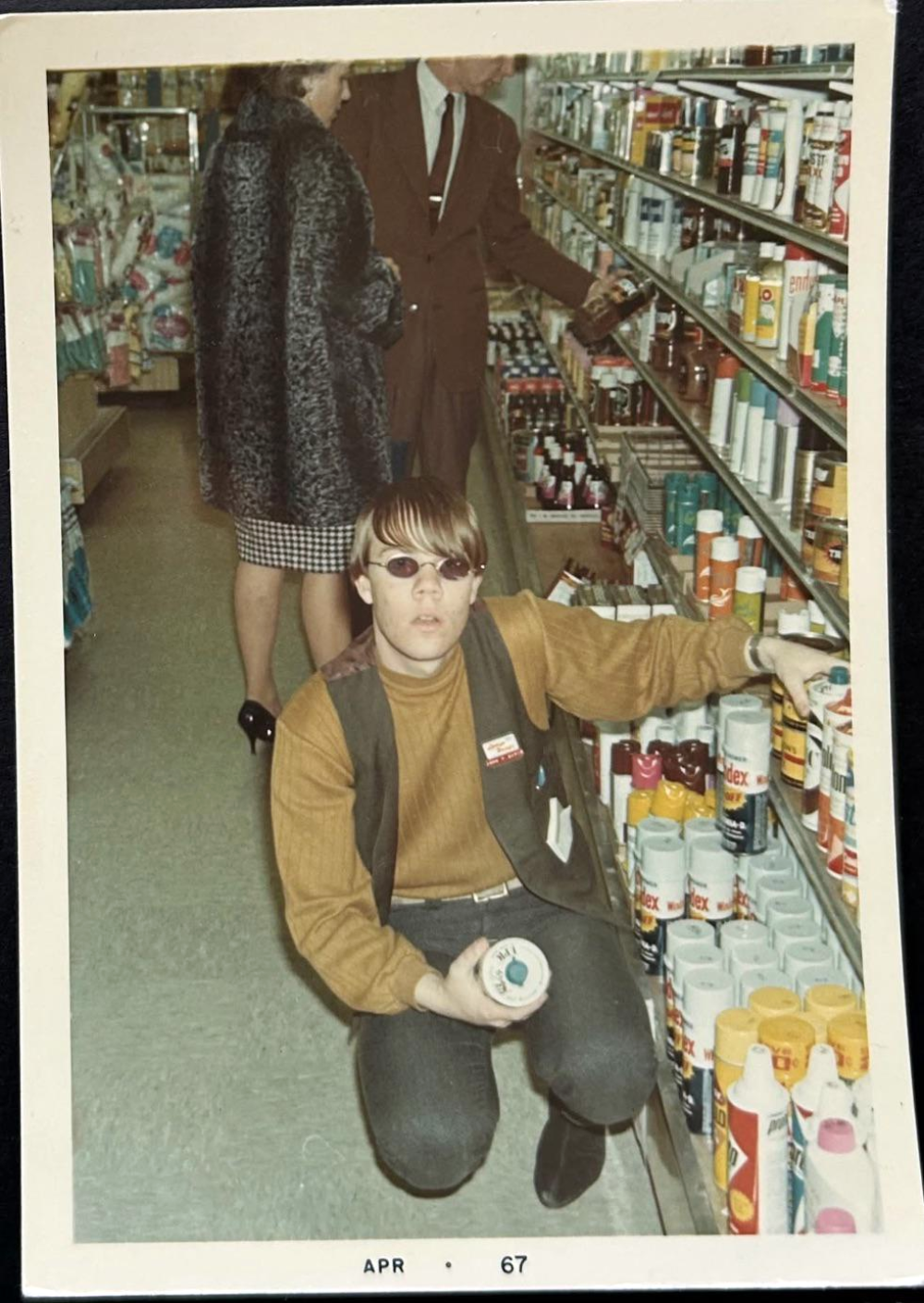 A young man wearing sunglasses crouches in an aisle of a store holding a can of spray paint, while a person in a coat stands in the background looking at shelves.