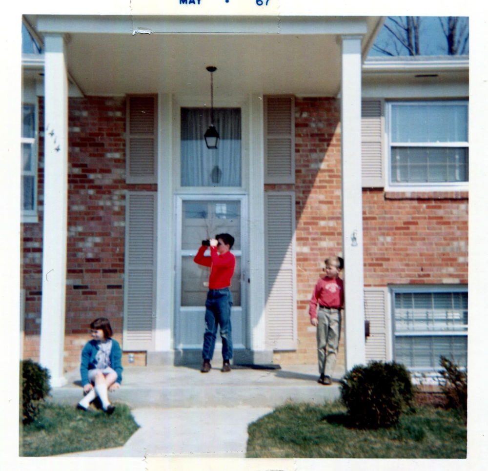 Two boys and a girl standing on the front porch of a brick house, one boy looking through binoculars.