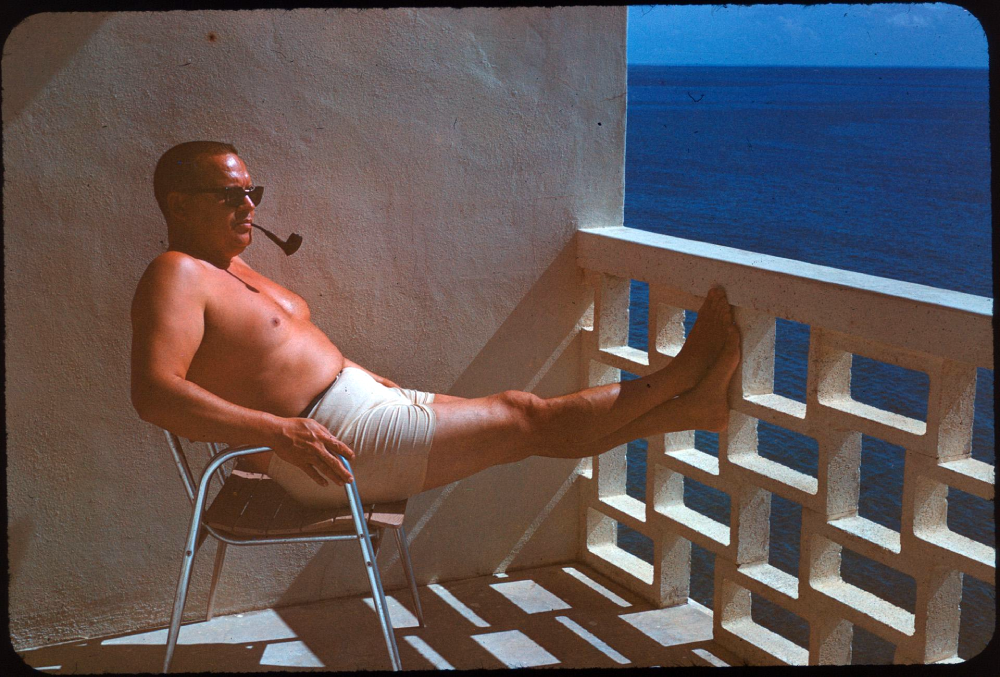 A shirtless man in sunglasses and shorts sits in a chair on a balcony, smoking a pipe with his feet propped up against a decorative railing overlooking the ocean.