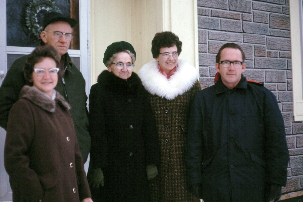 Five people stand in a row outside a house, wearing winter coats.