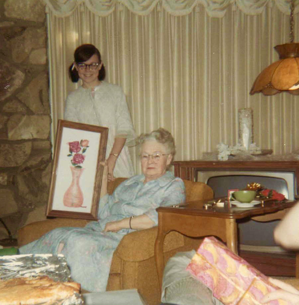 A young girl stands behind an older woman seated in an armchair, holding up a framed floral painting for her to see.