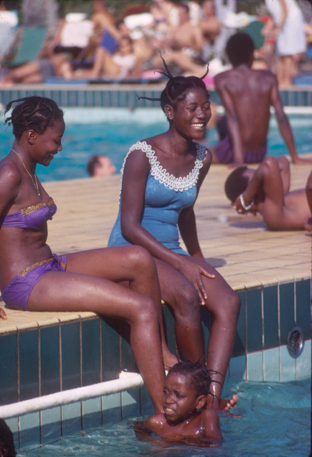 Two women in swimsuits sit on the edge of a swimming pool, laughing, while a child plays in the water in front of them.