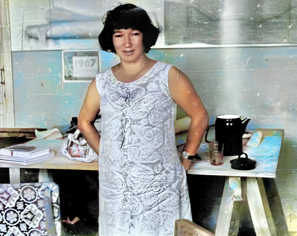 A woman with short dark hair stands in front of a desk wearing a sleeveless white patterned dress.