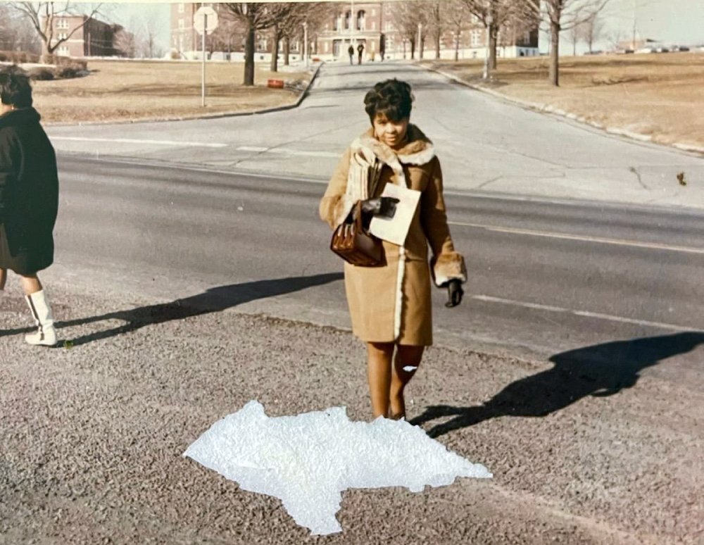 A woman wearing a tan coat and gloves holds a folder while walking across a paved area near a road.