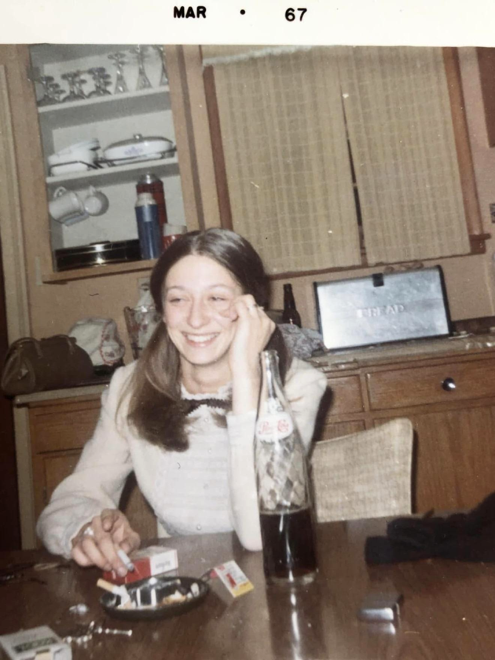 A smiling woman sits at a table with a glass soda bottle, a pack of cigarettes, and an ashtray, in a kitchen dated March 1967.