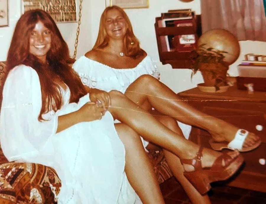 Two women with long hair and white dresses sit indoors, smiling. One is seated on a chair and the other on a desk, with a globe, books, and a plant in the background. Both have crossed legs and are wearing sandals.