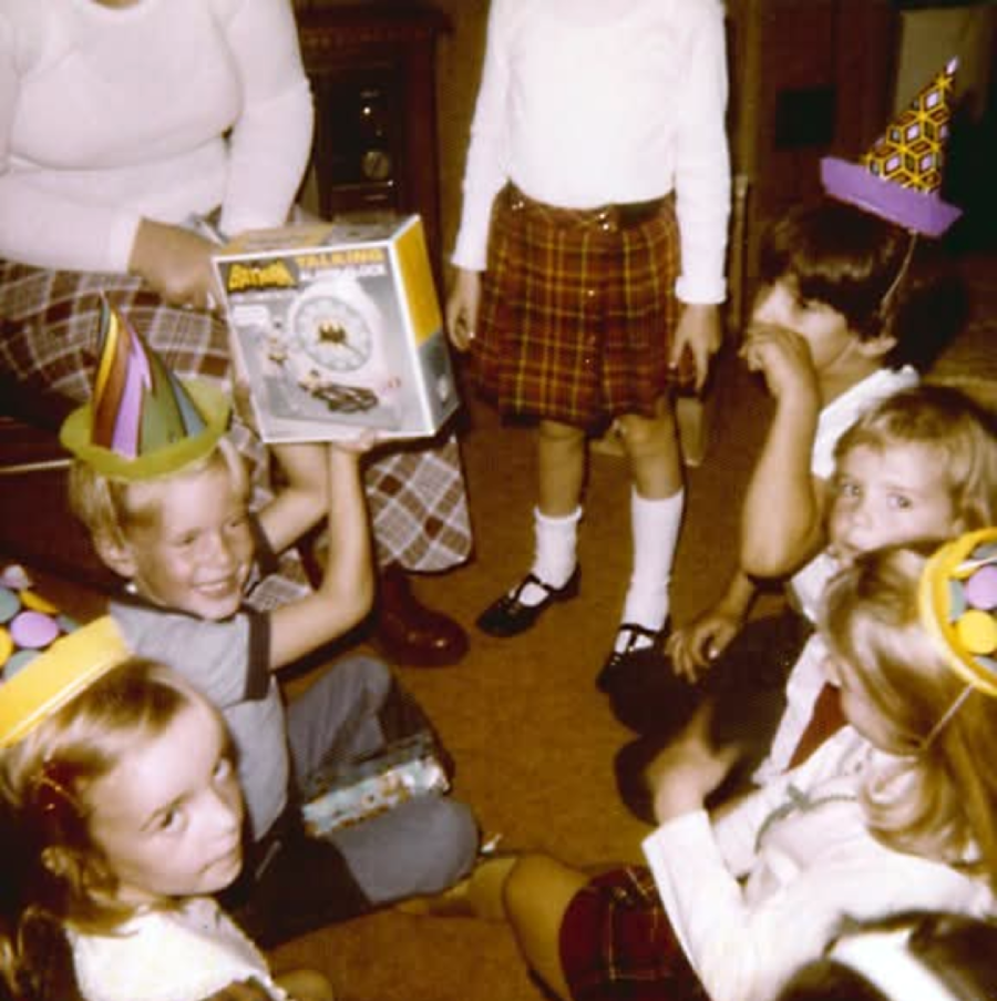 Children in party hats sit in a circle on the floor, one child holds up a toy box. An adult and another child stand nearby. The group appears to be celebrating a birthday in a cozy indoor setting.