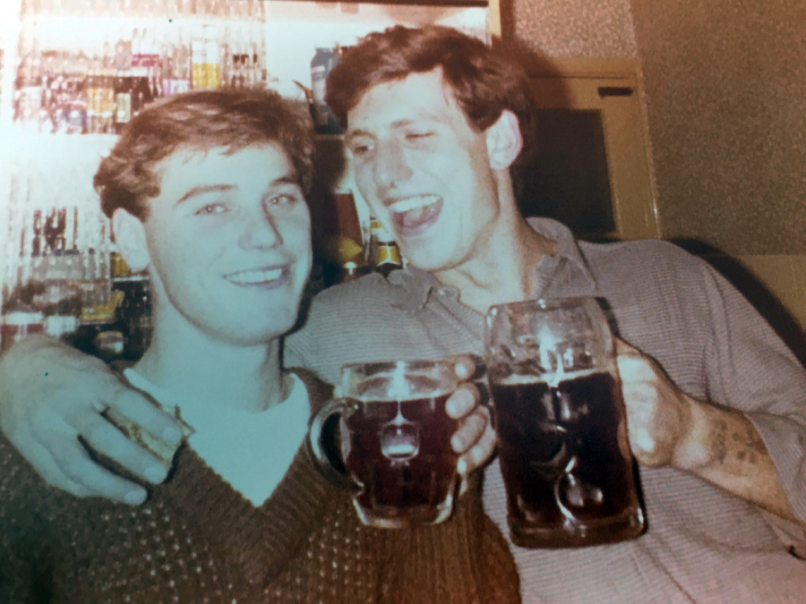 Two young men with short brown hair smile and pose together, each holding a large beer mug. One man has his arm around the other’s shoulder. Shelves with drinks are visible in the blurry background.