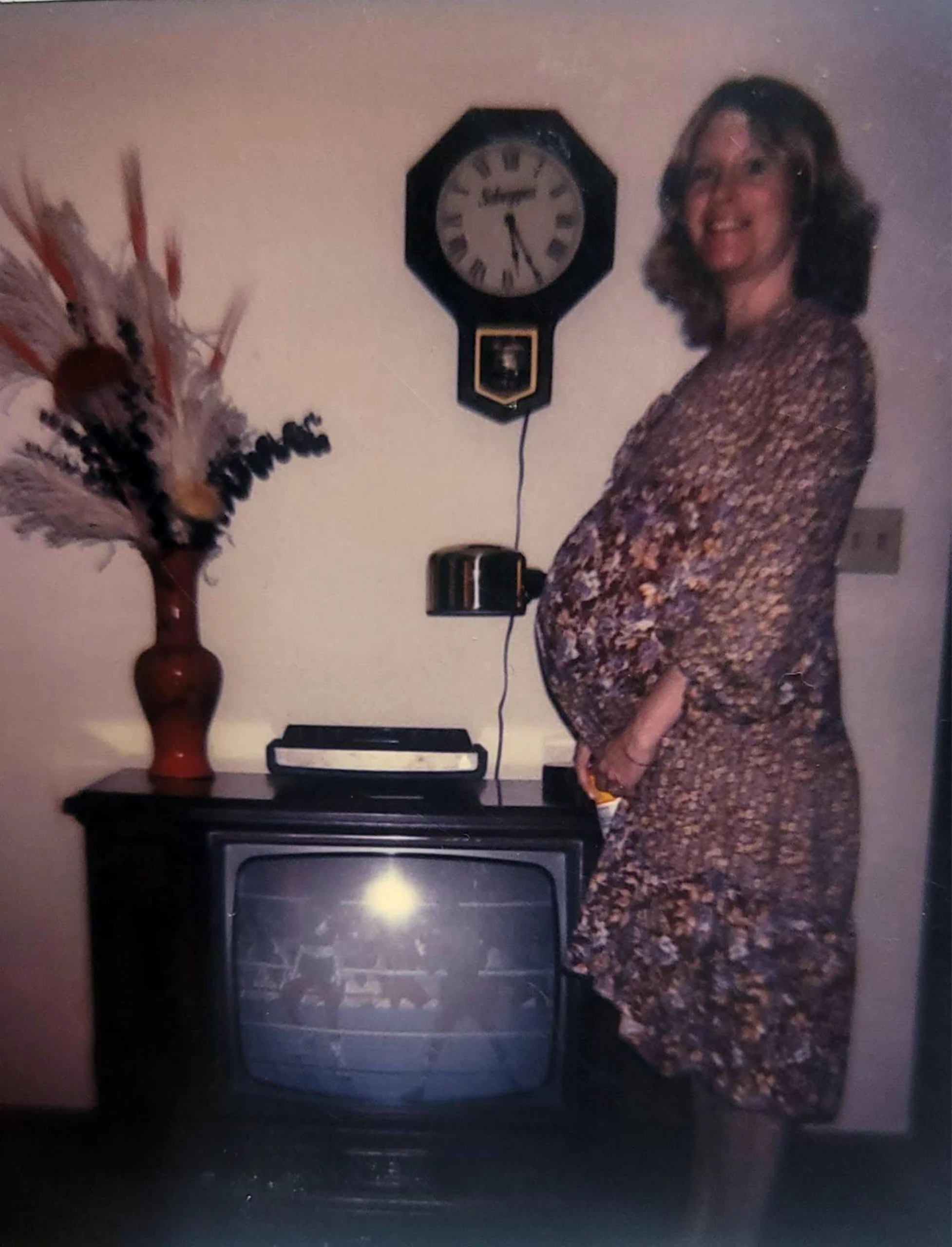 A pregnant woman in a floral dress stands by an old TV and a vase of dried flowers. A wall clock and a small shelf are above the TV. The woman is smiling and looking at the camera.