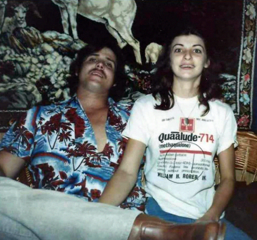 A man in a blue Hawaiian shirt and a woman in a Quaalude T-shirt sit closely together on a couch, holding hands, in front of a tapestry with animal designs.