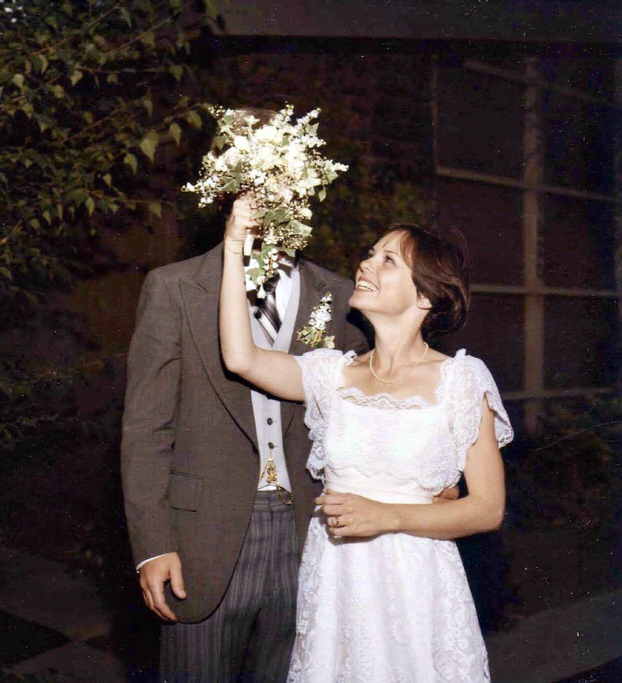 A bride in a white dress playfully covers the groom’s face with her bouquet while smiling, as he stands beside her in a brown suit.