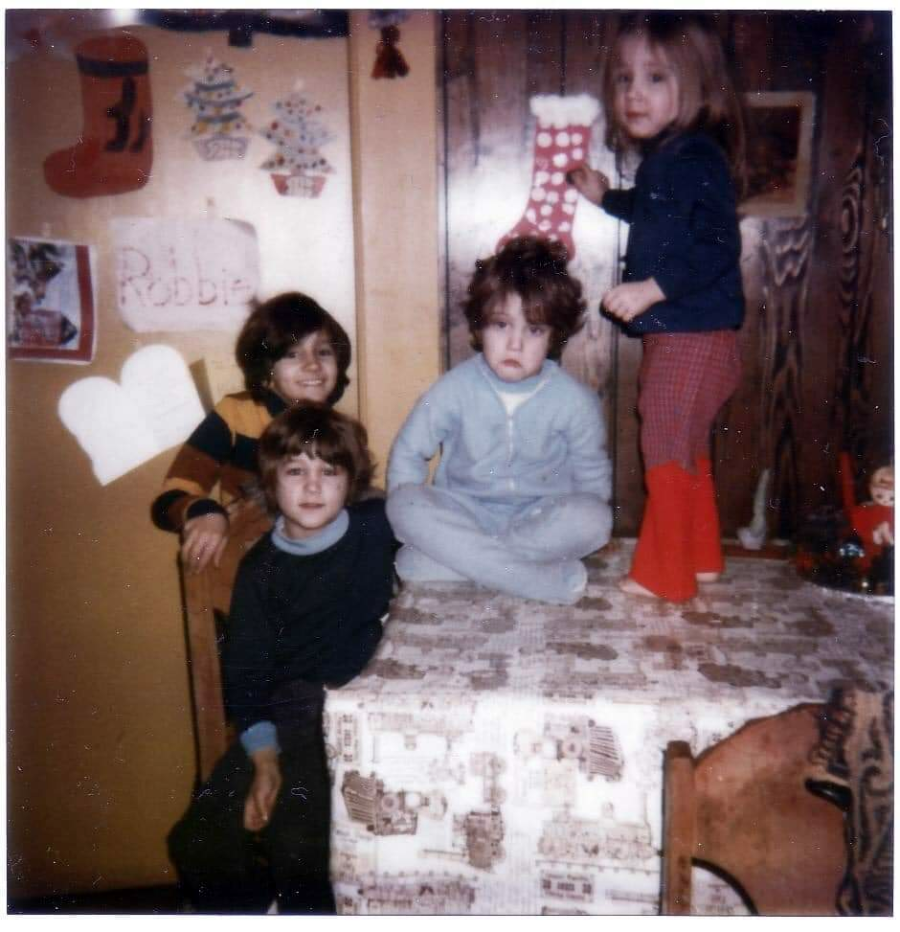 Four children pose indoors, three sitting on a table and one standing, surrounded by holiday decorations including stockings and festive cutouts on the wall. A handwritten sign says “Robbie.”