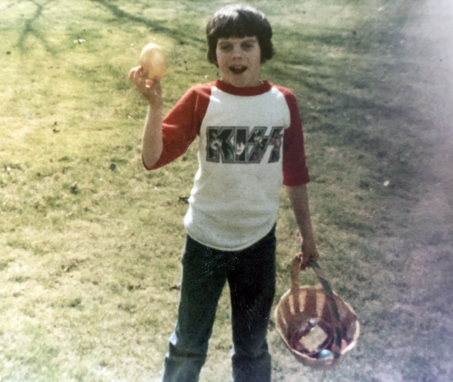 A child stands on grass holding a basket and a large plastic egg, wearing a red and white KISS shirt, smiling outdoors.