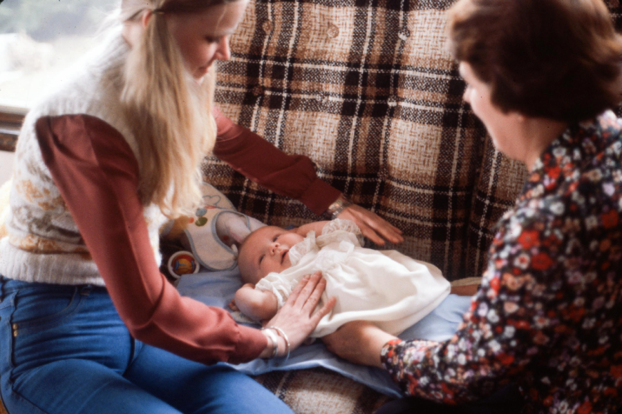 Two women sit on a plaid couch and gently touch a baby dressed in white, who is lying on a blue blanket. The scene appears warm and caring, with both women focusing on the baby.
