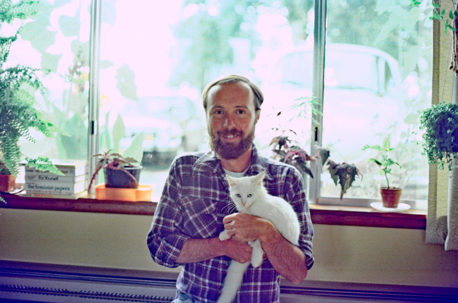 A man with a beard and plaid shirt holds a white cat in front of a window with potted plants. Sunlight streams in from outside, creating a bright, relaxed scene.