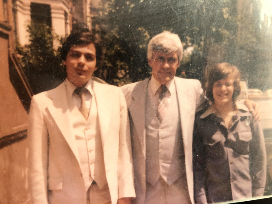 Three people pose outdoors. Two adults in light-colored suits stand next to a smiling young person in a dark shirt. Trees and a building are visible in the background. The image has a warm, vintage look.