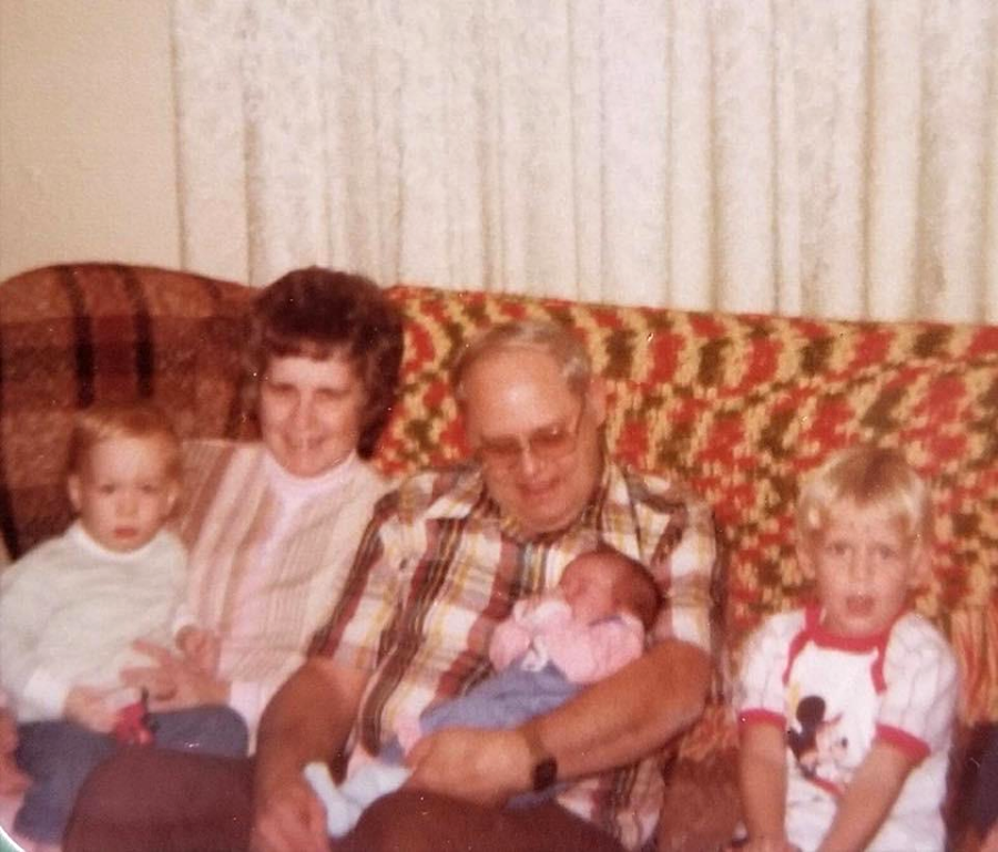 An elderly couple sits on a couch with three young children; the man holds a baby, and two toddlers sit on either side. A colorful afghan blanket is draped on the couch.