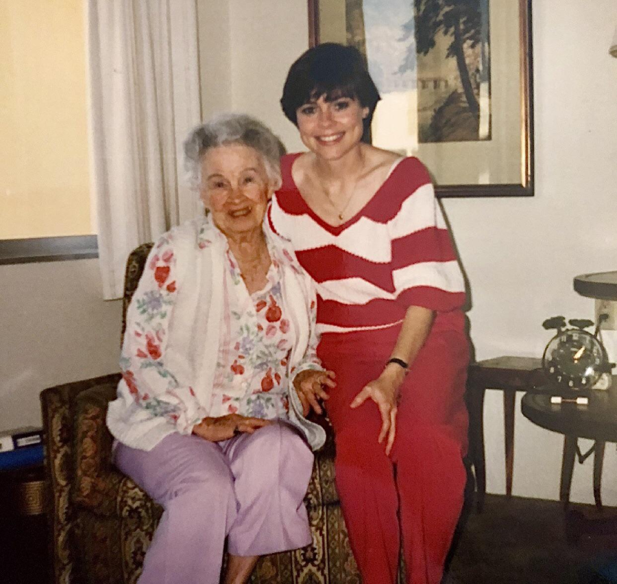 Two women sit together indoors, smiling. The older woman wears a floral blouse and lilac pants, the younger wears a red and white striped sweater with red pants. There’s a framed picture and a rotary phone in the background.