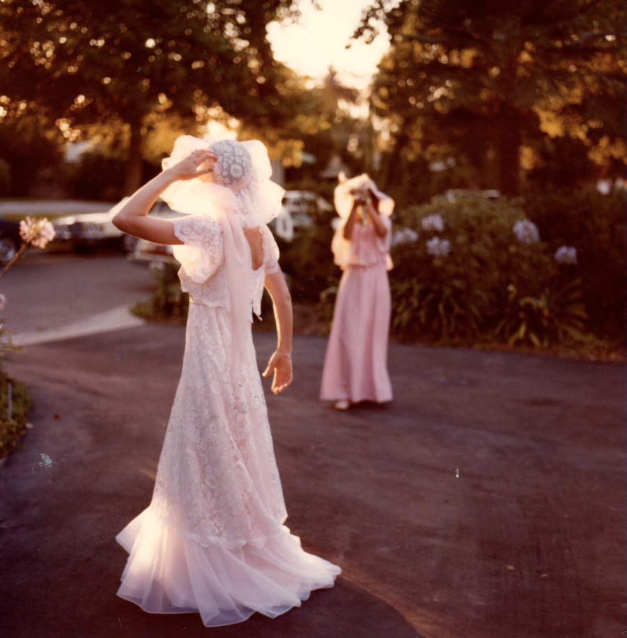 Two women in vintage dresses and hats stand on a driveway at sunset. One wears white lace, the other wears pink. Trees, flowers, and classic cars are visible in the background, bathed in warm sunlight.